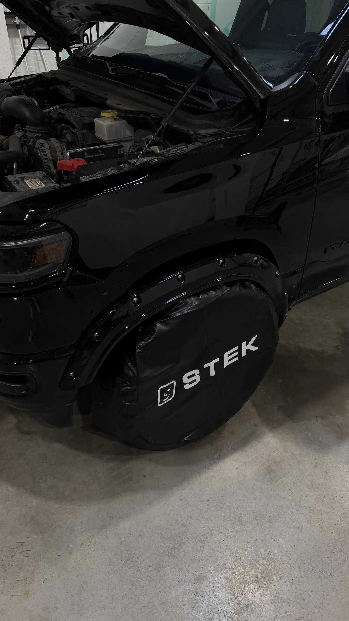 A black RAM 1500 in the STEK Estonia workshop, hood up, with gloss black bolt-on fender flares around the front wheel arch.